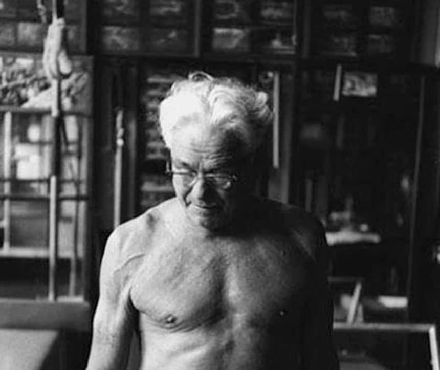 Joseph Pilates em seu estúdio, já idoso e sem camisa, demonstrando vitalidade e força física, cercado por aparelhos do método que criou.