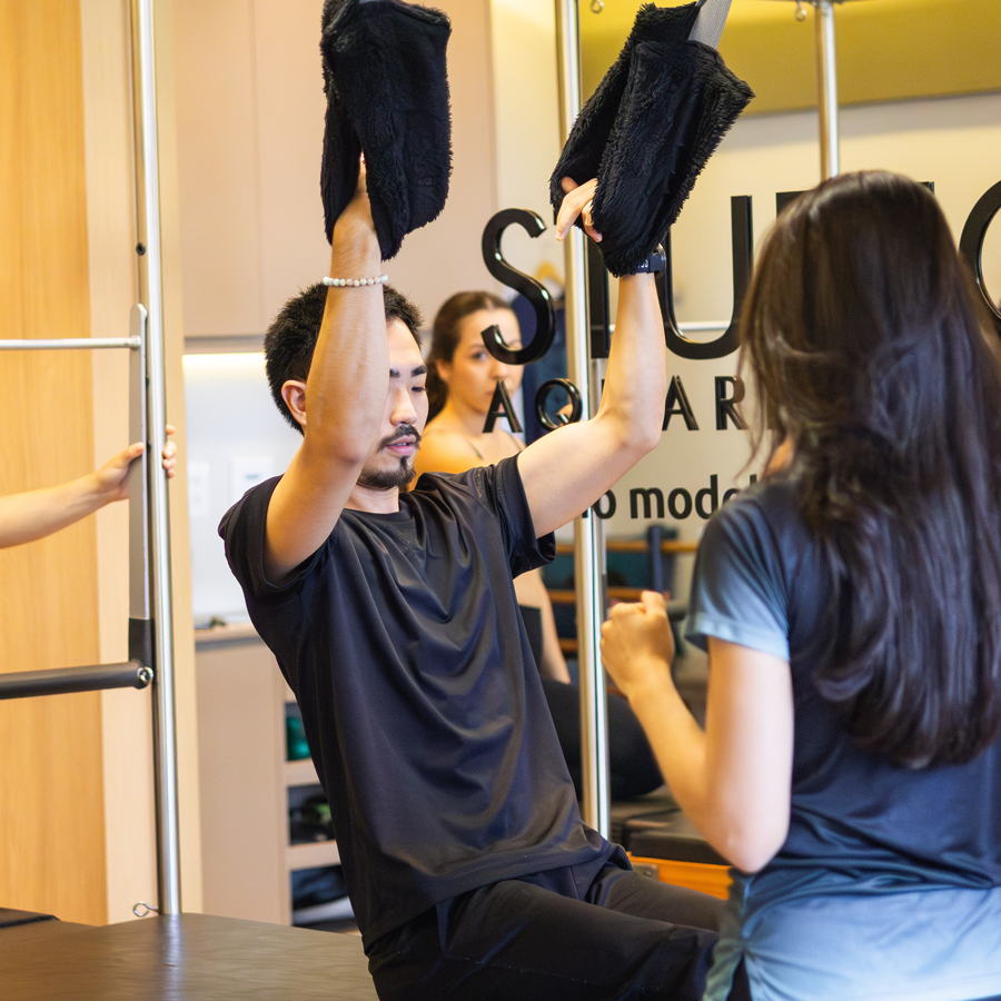 Homem realiza exercício no Cadillac com auxílio de alças, sob orientação de uma instrutora de pilates dentro de um estúdio moderno.